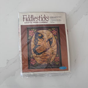 May include: A Fiddlesticks Crafts kit featuring a stained glass butterfly long stitch picture. The kit includes a canvas, yarn, needle, and instructions. The butterfly design is in shades of gold, blue, and brown, surrounded by pink flowers.
