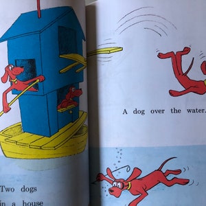 May include: A children's book illustration shows red dogs in various scenes. One dog rows a boat with a blue house, while another jumps over water. The text reads, "Two dogs in a house" and "A dog over the water."