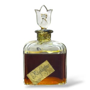 May include: A vintage perfume bottle with a clear, square-shaped glass body. The bottle contains a dark amber liquid and features a decorative stopper with a white tulip design and the letter "R". A gold label reads "Replique Paris".