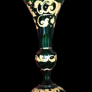 Puede incluir: Un jarrón alto de cristal verde oscuro con la parte superior acampanada y una base ancha. El jarrón está adornado con intrincados diseños florales y de volutas de color dorado. El jarrón tiene una sección media bulbosa y un cuello delgado.