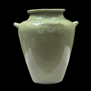 Könnte beinhalten: Eine hellgrüne Keramikvase mit breitem Körper und schmalem Hals. Die Vase hat zwei kleine Griffe an den Seiten und eine leicht strukturierte Oberfläche. Die Vase steht vor einem schwarzen Hintergrund.