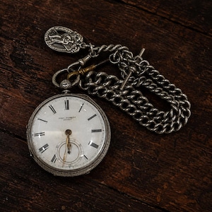 May include: A silver pocket watch with a white face, black Roman numerals, and the text John Forrest London. It features a silver chain with a T-bar and a circular silver pendant depicting a figure carrying a cross, attached to the watch bow.