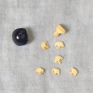 May include: A selection of small, ivory-coloured elephant figurines and a dark, round object, possibly a bead or a button, are arranged on a light grey fabric. The elephants are intricately carved, showcasing their forms. The objects are scattered across the fabric.
