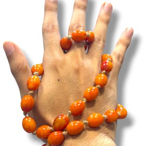 Könnte beinhalten: Ein Halsketten- und Armband-Set mit ovalen, orangefarbenen Perlen, die auf einer naturfarbenen Kordel aufgereiht sind. Die Perlen haben eine glänzende Oberfläche und variieren leicht in der Farbe, wobei einige rote Farbtöne aufweisen. Der Schmuck wird auf einer Hand vor weißem Hintergrund präsentiert.