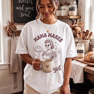 Könnte beinhalten: Ein weißes T-Shirt mit einer braunen Grafik einer Person mit Kopftuch, die ein Brot hält, mit dem Text MAMA MAKES und dem unten teilweise sichtbaren Wort dough. Das Design enthält kleine Sternakzente und ein kariertes Schürzendetail.