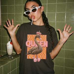 Könnte beinhalten: Ein dunkelgraues T-Shirt mit einem Grafikdruck eines Schwans im Leopardenmuster, der eine Krone und flauschige rosa High-Heels trägt. Das Wort QUEEN ist in großen, kräftigen, hellrosa Buchstaben auf einem orangefarbenen, welligen Hintergrund gedruckt.