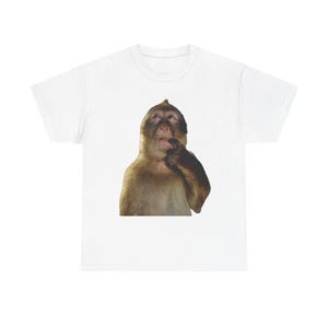 Peut inclure: T-shirt blanc avec un dessin de singe pensif, touchant son menton. Le singe a des poils bruns et beiges. Le t-shirt est de style col rond.