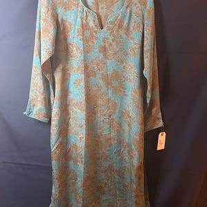 May include: A long-sleeved tunic featuring a light blue and tan floral pattern, a notched neckline, and a small tag attached to the sleeve with the text 36 inches. The garment has a straight hem and a relaxed fit throughout the body.