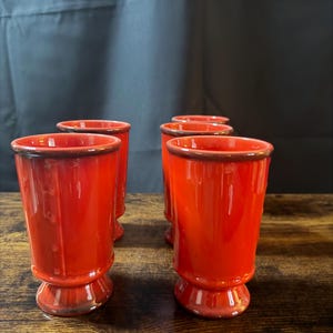 May include: A set of four vibrant orange ceramic tumblers with a glossy finish and a dark brown rim. Each tumbler features a cylindrical body that tapers slightly toward a circular pedestal base, creating a uniform look across the collection.