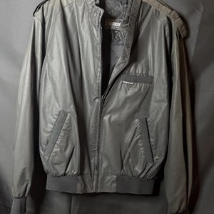 May include: A black Members Only jacket featuring a front zipper closure, a stand-up collar with a snap button tab, epaulets on the shoulders, two side pockets, and a chest pocket with the Members Only logo tag. The jacket has ribbed cuffs and hem.