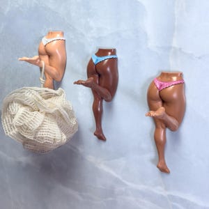 Puede incluir: Tres esculturas murales de piernas y glúteos humanos en varios tonos de piel, cada una con una tanga de diferente color. Una escultura sostiene una esponja de baño de malla blanca.