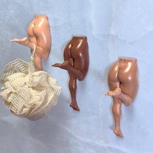 Puede incluir: Tres esculturas murales de piernas y glúteos humanos en diferentes tonos de piel. Una sostiene una esponja vegetal beige. Las esculturas están hechas de un material liso y brillante y están montadas sobre un fondo azul claro.