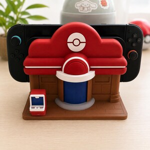 Op de afbeelding: Een dockingstation voor een spelcomputer in de vorm van een rood, wit en bruin gebouw met een centraal Poké Ball-embleem. De structuur heeft een blauwe deur, een kleine rood-witte accessoirestandaard en houdt een zwarte handheld-console vast.