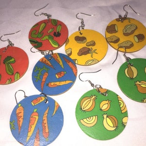 May include: A set of eight colorful round earrings with different vegetable designs. The earrings are made of wood and have silver hooks. The designs include carrots, onions, potatoes, and celery.