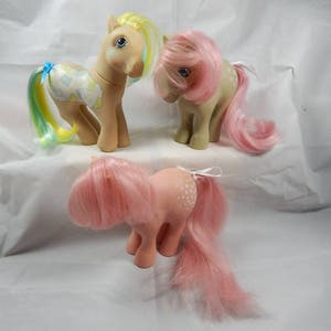 May include: Three vintage My Little Pony toys. The ponies are all standing on a white surface. The pony on the left has a yellow and blue mane and tail, and a blue and white patterned body. The pony in the middle has a pink mane and tail and a beige body. The pony on the right has a pink mane and tail and a pink body with white dots.