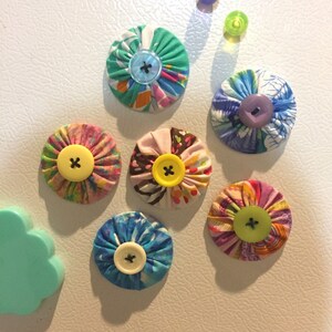 May include: Six colorful fabric flower magnets with different colored buttons in the center. The magnets are attached to a white refrigerator.