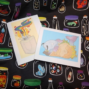 May include: Two greeting cards with colorful illustrations. The first card features a rainbow-colored smoke coming out of a pocket with a lighter. The second card shows a fish in a taco shell.