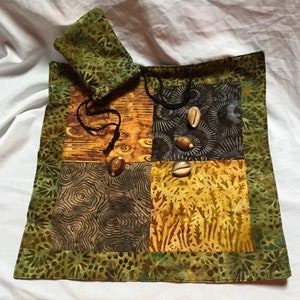 May include: A green fabric pouch with a drawstring closure, sitting on a square patchwork fabric mat with a green border. The mat is made of four different patterned fabrics in shades of brown, yellow, and black. There are four small, light brown seashells on the mat.
