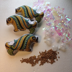 May include: Three plush toys shaped like fish with a colorful, striped pattern in shades of yellow, green, and blue. The toys are on a white surface with a pile of brown cat food pellets.