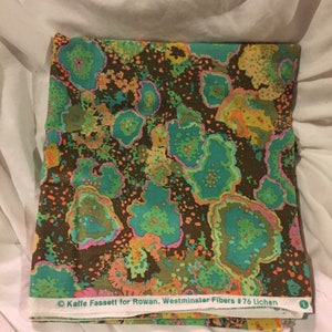 May include: A folded piece of fabric with a brown background and a colorful abstract pattern of green, pink, orange, and yellow. The fabric has a label that reads "Kaffie Fassett for Rowan. Westminster Fibers #76 Lichen."