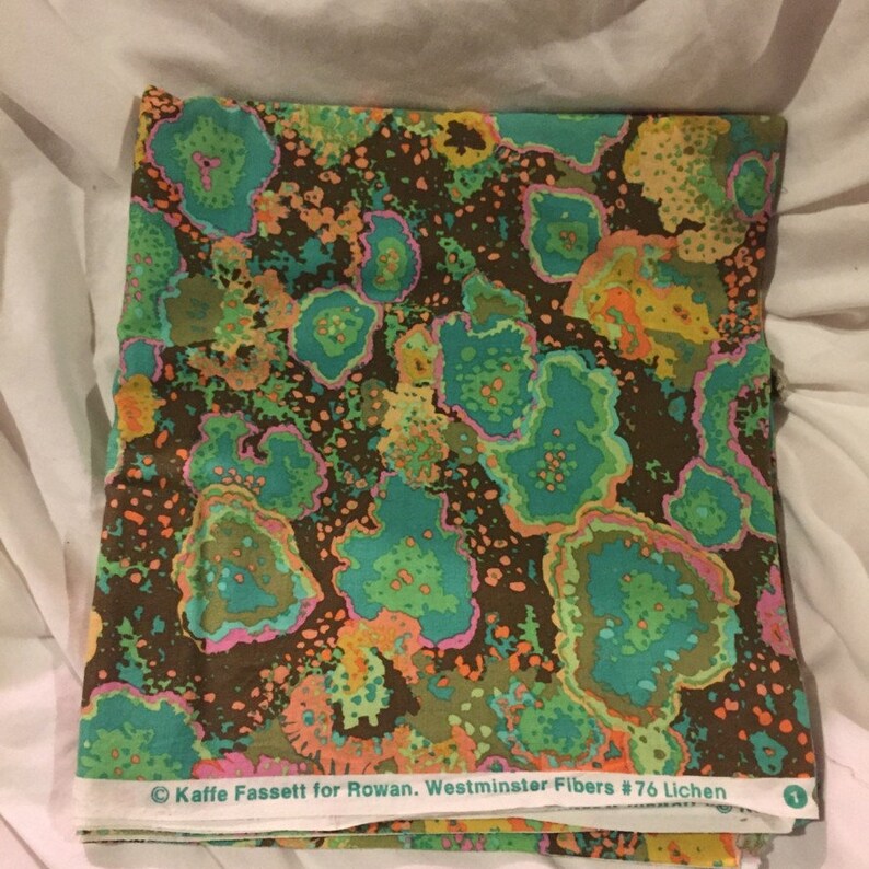 May include: A folded piece of fabric with a brown background and a colorful abstract pattern of green, pink, orange, and yellow. The fabric has a label that reads "Kaffie Fassett for Rowan. Westminster Fibers #76 Lichen."