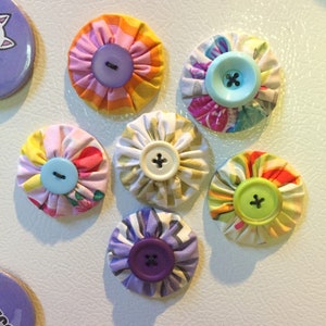 May include: Six colorful fabric covered button magnets with different patterns and colors. The magnets are round and have a button in the center. The buttons are different colors, including purple, green, blue, and white.