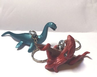 May include: Two keychains featuring a blue plesiosaur and a red octopus. The plesiosaur is a long-necked marine reptile with flippers and a long tail. The octopus has eight tentacles and is curled up in a defensive position. Both keychains are made of plastic and have silver metal key rings.