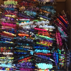 May include: A box filled with colorful yarn skeins, each wrapped in brown paper and tied with a string. The yarn is a variety of textures and colors, including pink, blue, green, purple, and orange.
