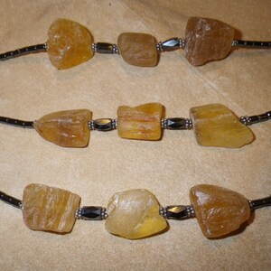 May include: Three strands of necklaces with rough-cut, amber-colored stones. The stones are interspersed with small, dark beads and metallic accents. The necklaces are displayed against a neutral, tan background.