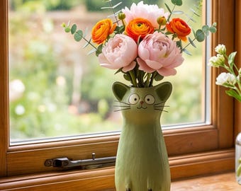 May include: A matte olive green ceramic vase shaped like a cat with a small face, whiskers, and pointed ears. The vase holds a bouquet of pink peonies, orange ranunculus, and green eucalyptus stems. A clear glass vase with white flowers sits nearby.