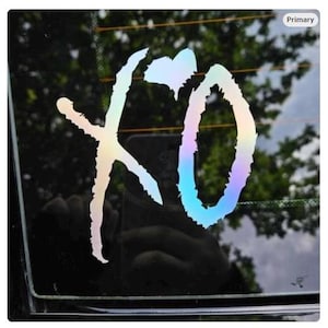 May include: Holographic sticker featuring the letters "XO" in a stylized script. The sticker has a rainbow iridescent finish, reflecting various colors. The sticker is affixed to a car window, with a blurred background of trees.