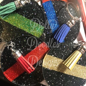 May include: Four circular black glitter keychains, each featuring a horizontal glitter stripe in green, blue, red, or gold. Each keychain includes a matching colored suede tassel attached to a silver-toned metal ring and hardware.