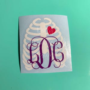 May include: A white vinyl decal featuring a stylized rib cage design with a red heart and a monogram in purple. The monogram includes the letters "LOL" in a decorative font, set against a light blue background.