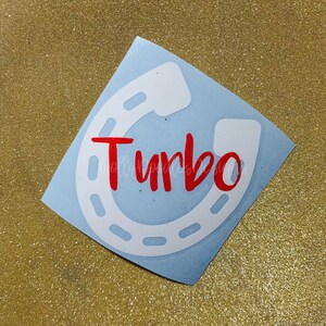 May include: A light blue sticker featuring a white horseshoe design with the word "Turbo" in red cursive script. The sticker is on a gold glitter background.
