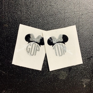 May include: Two white square stickers featuring a black Minnie Mouse silhouette with a silver glitter bow. The center of each sticker has a silver glitter monogram. One sticker reads "GOP" and the other reads "10".