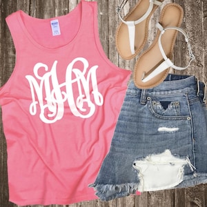 May include: A pink tank top with a white monogram, paired with denim shorts and white sandals. The shorts have distressed details. The sandals have a neutral sole and white straps. The outfit is displayed on a wooden surface.