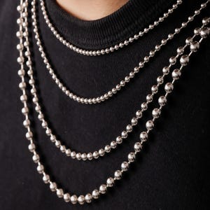 May include: A set of four silver-toned ball chain necklaces of varying lengths and bead sizes. Each necklace consists of small, polished spherical metal beads connected in a continuous line, draped in layers against a plain black fabric background.