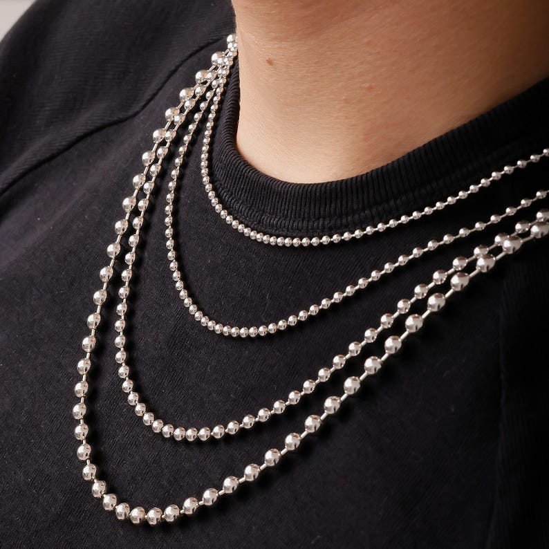 May include: A set of three layered silver-toned ball chain necklaces. Each necklace features a series of small, reflective, spherical metal beads connected in a continuous strand, creating a textured, metallic appearance against a black shirt.