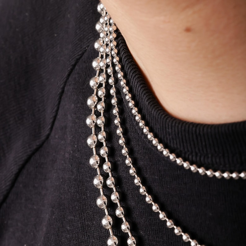 May include: A layered necklace set consisting of four distinct silver-toned ball chains. The chains feature varying bead sizes, ranging from small, uniform spheres to larger, graduated metallic balls, all resting against a black fabric background.