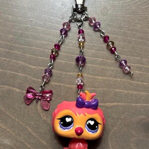 May include: A handmade keychain featuring a small, colorful owl figurine with a purple bow. The keychain includes a silver clasp, beaded chains with pink and purple beads, and a pink bow charm. The owl is orange and pink.