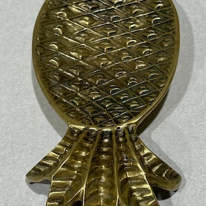 May include: A polished brass decorative tray shaped like a pineapple, featuring a textured diamond-patterned body and a detailed leafy top section. The metallic surface reflects light, highlighting the intricate grooves and embossed pineapple design.