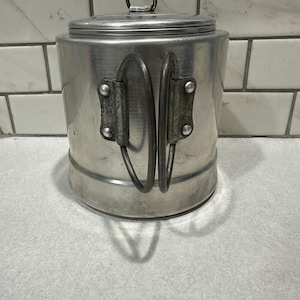 May include: A cylindrical silver-colored metal canister with a flat lid and a small circular handle on top. The container features two vertical, hinged wire handles attached to the side with metal rivets, providing a sturdy grip for the vessel.