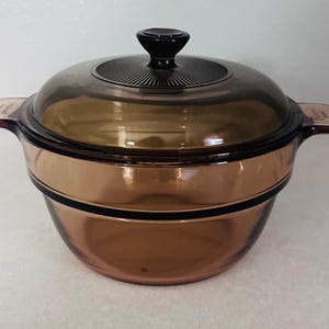 May include: A brown glass casserole dish with a lid. The dish has a round shape with two handles and a black rim. The lid has a black knob and a ridged design. The dish is made of a transparent, heat-resistant glass.