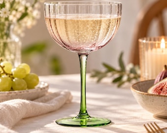 May include: A stemmed coupe glass featuring a ribbed, light pink bowl filled with a sparkling golden liquid. The glass has a slender, vibrant green stem and a matching green circular base, creating a distinct contrast between the pink and green.