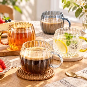 May include: Four round, ribbed glass mugs with handles, each containing a different beverage. One mug holds dark coffee, another amber tea, a third a light brown drink, and the fourth a clear sparkling liquid with a lemon slice and mint leaves.