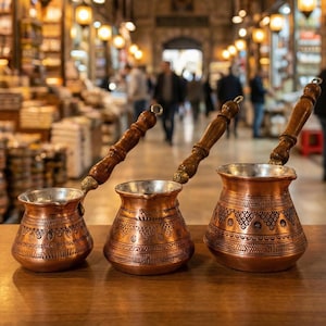 May include: Three copper coffee pots with wooden handles are displayed on a wooden surface. The pots have intricate etched designs and a shiny interior. The background shows a bustling marketplace with people and warm lighting.