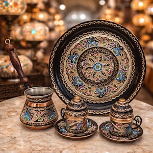 Op de afbeelding: Een sierlijke Turkse koffieset met een koperen cezve, twee kleine kopjes met schotels en een grote serveerschaal. De set is zwart met gouden en kleurrijke bloemmotieven. De cezve heeft een houten handvat.