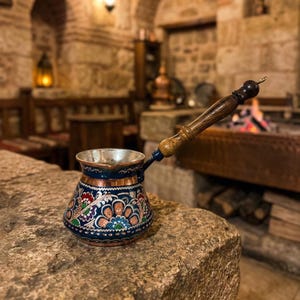 May include: A handcrafted copper coffee pot, featuring a dark blue base with intricate floral patterns in red, white, and green. The pot has a wooden handle and sits on a stone surface, suggesting a traditional setting.