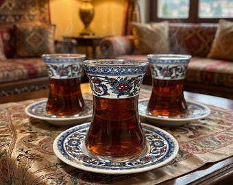 以下が含まれることがあります： 琥珀色の紅茶が入った3つの装飾的なトルコ風ティーグラス。お揃いのソーサーの上に置かれています。グラスとソーサーには、青、赤、白の花柄模様が施されています。模様入りのテーブルクロスと濃い木製のテーブルの上に配置されています。