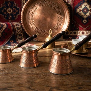 Puede incluir: Tres cafeteras de cobre con mangos de madera sobre una mesa de madera. Las ollas tienen patrones intrincados y están acompañadas de bandejas de cobre, tazas y un plato decorativo. El fondo presenta alfombras estampadas y un plato de cobre.